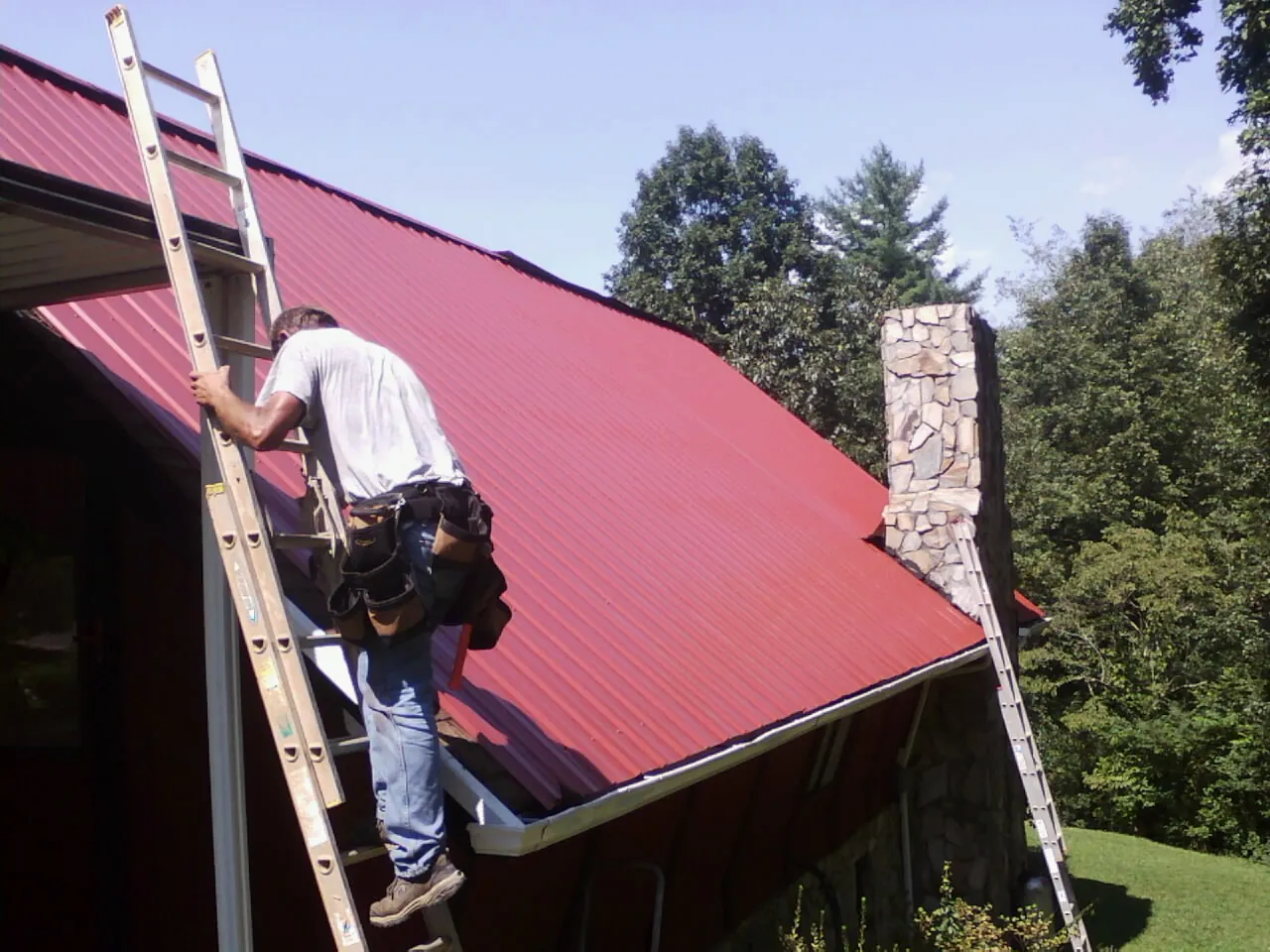 Roofer on a ladder beside a red metal roof and stone chimney — Soffit & Fascia Repair by Roofing Brothers in Diggs
